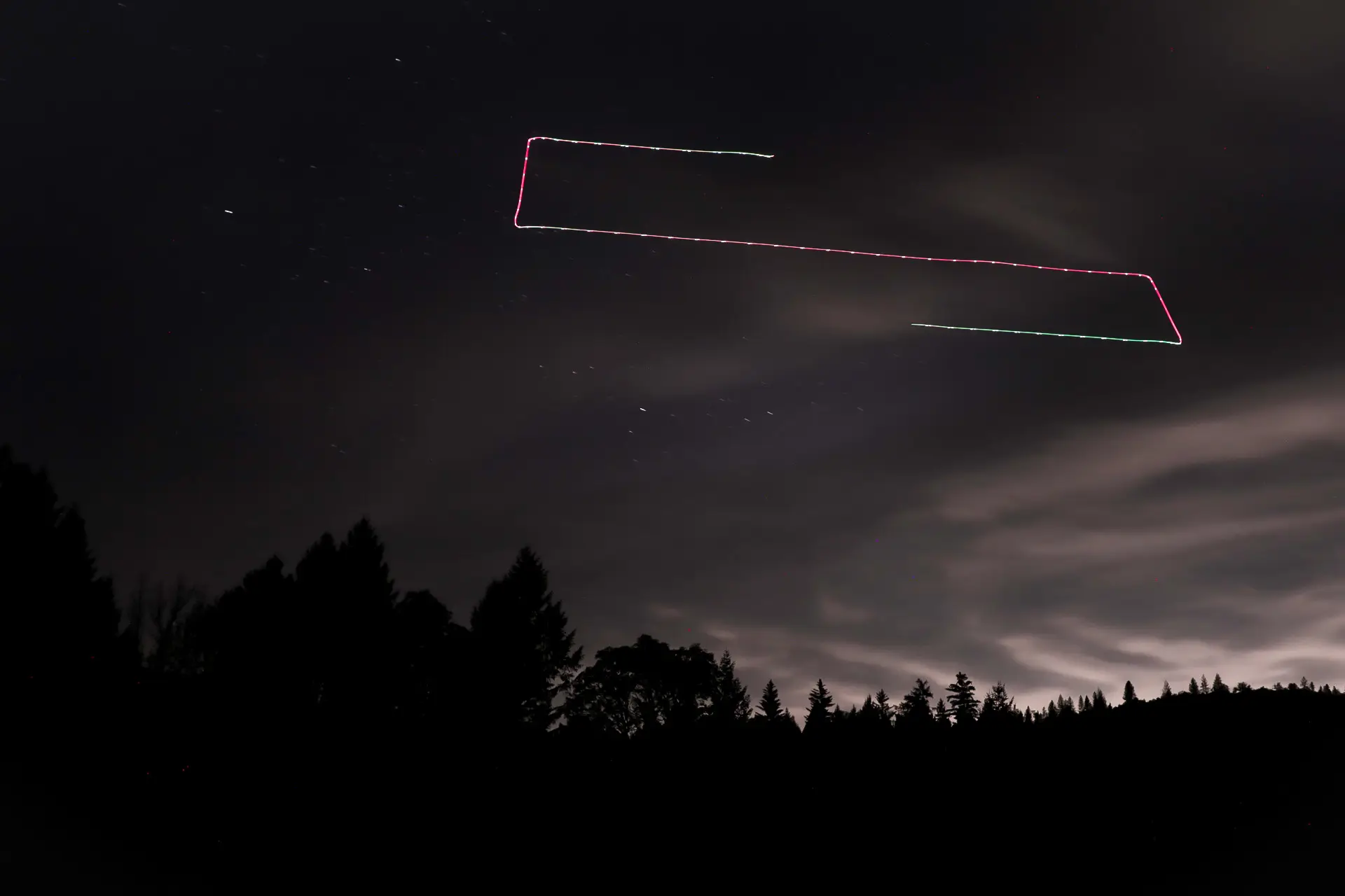 Long exposure photograph of a thermal drone gridding the night sky in a rectangular survey pattern over a forest silhouette.